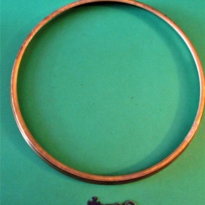 Puede incluir: Un aro de metal redondo de color cobre con una pequeña llave plateada con cabeza en forma de cruz que se encuentra debajo sobre una superficie verde.