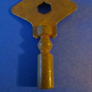 May include: A rusty metal key with a diamond-shaped head and a hexagonal hole in the center. The key has a round knob and a cylindrical shaft.
