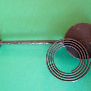 May include: A close-up of a clock spring and winding key. The spring is coiled and attached to a round metal piece. The winding key is attached to the spring and has a metal knob.