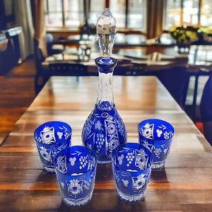 May include: A set of four blue crystal glasses and a matching decanter with intricate etched designs. The glasses and decanter are on a wooden table.