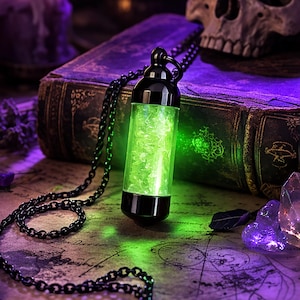 Peut inclure: Un collier à chaîne noire avec un pendentif vert brillant, sur fond mystique. La scène comprend un crâne, des bougies, un vieux livre et des cristaux, créant un thème gothique ou fantastique. Le pendentif est cylindrique avec des embouts noirs.