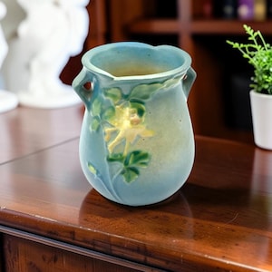 May include: A matte blue ceramic vase with two small handles at the rim. The exterior features a raised relief design of a yellow flower with green leaves. The vase has a rounded body and a wide, open mouth with a slightly scalloped edge.