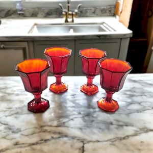 May include: Four red glass goblets with a clear amber tint and a faceted design. The goblets are arranged on a white marble surface.