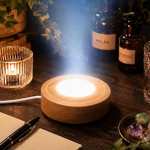 Puede incluir: Una base de exhibición LED redonda de madera clara con un cable de alimentación blanco. El centro de la base circular emite una luz blanca cálida y brillante que se proyecta hacia arriba. La base está sobre una superficie oscura junto a un cuaderno y un bolígrafo.