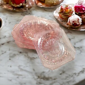 May include: A stack of four pink glass dessert plates with a delicate floral pattern. The plates have a vintage look and are perfect for serving small desserts or appetizers.