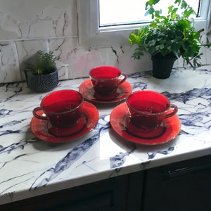 May include: Three red glass teacups and saucers with a gold rim. The cups and saucers are on a white marble countertop.