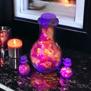 May include: Three glass bottles with cork stoppers filled with colorful, translucent, and iridescent stones. The bottles are illuminated from within, casting a warm glow. The bottles are on a black surface.