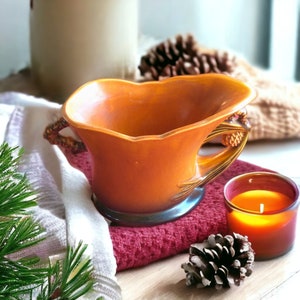 May include: An orange ceramic gravy boat with a decorative handle featuring a pinecone design. The gravy boat sits on a red towel, next to a lit candle and a pinecone. A sprig of pine and a white towel are also in the image.