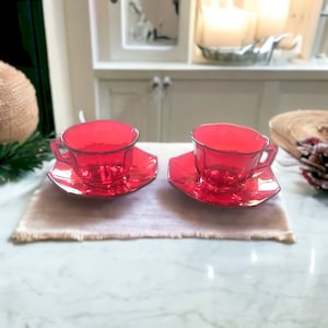 May include: Two red glass teacups and saucers. The cups and saucers are a deep red color and have a shiny finish.