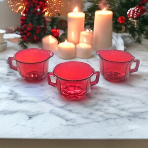 May include: Three red glass creamer pitchers on a white marble surface. The pitchers have a rounded shape with a small spout and two handles.