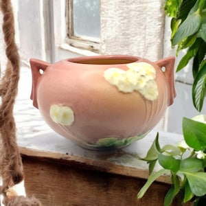 May include: A pink ceramic bowl with a floral design. The bowl has two handles and is decorated with white and yellow flowers and green leaves. The interior of the bowl is a light yellow color.