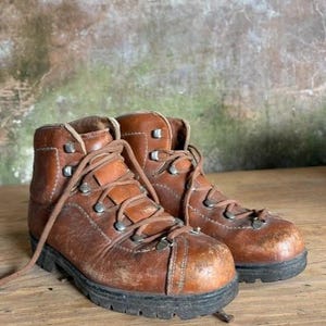 May include: A pair of brown leather hiking boots featuring a high-top design, metal eyelets for the laces, and thick black rubber soles. The leather shows signs of wear with scuffs on the rounded toe areas and visible stitching details.