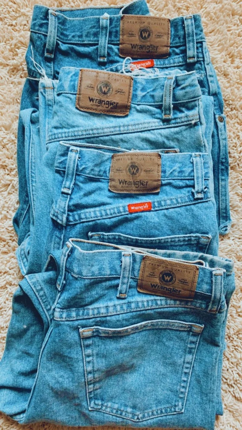 May include: Four pairs of blue denim jeans stacked on top of each other. Each pair has a brown Wrangler label with a white 'W' logo and the word 'Wrangler' printed in white.