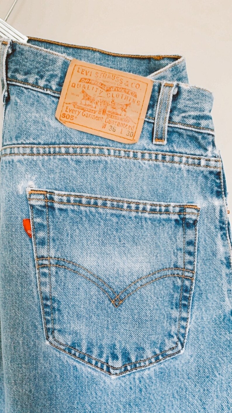 May include: A pair of light blue denim jeans with a brown leather Levi Strauss & Co. patch that reads "Quality Clothing" and "Every Garment Guaranteed" with the size 36 and 30. The jeans have a back pocket with a red tab.