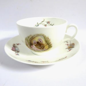 Peut inclure: Un ensemble tasse et soucoupe en céramique blanche orné d'illustrations de petites souris brunes dans diverses activités. La tasse montre une souris sortant d'un terrier, tandis que la soucoupe présente des souris avec des motifs floraux et des vignes.