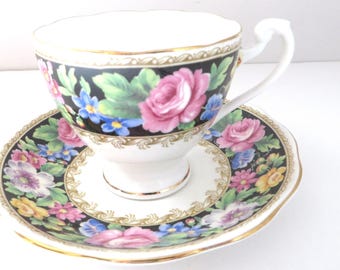 May include: A porcelain teacup and saucer set featuring a black background with a vibrant floral pattern of pink roses, blue forget-me-nots, and green leaves. The set is accented with gold trim along the scalloped edges and a decorative border.