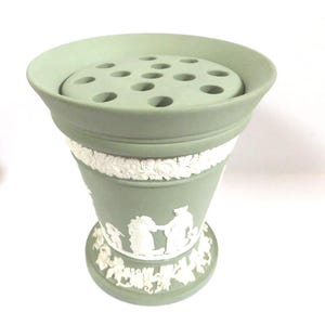 May include: A pale green ceramic flower frog vase with a wide, fluted top and a series of holes for arranging flowers. The vase features white relief detailing of classical figures and floral patterns around the body and base.