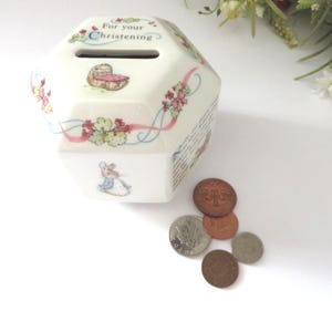 May include: A white ceramic money box with a hexagonal design, decorated with illustrations of a baby and a mouse. It features floral patterns and the text "For your Christening". Several coins are scattered nearby.
