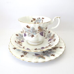 May include: A white porcelain teacup, saucer, and small plate set featuring a scalloped edge and gold trim. The pieces are decorated with a pattern of purple grape clusters and light blue and brown vine leaves against a white background.