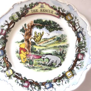 May include: A white ceramic decorative plate featuring a colorful illustration of Winnie the Pooh, Piglet, Eeyore, and Tigger. The center scene shows the characters holding a blanket, with the text THE RESCUE at the top and honey pots on the border.