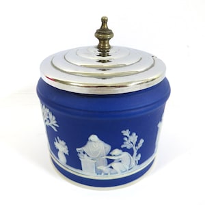 Peut inclure: Un pot cylindrique bleu à la finition mate orné de reliefs blancs représentant des personnages et des arbres. Le pot est doté d'un couvercle argenté poli à gradins avec un petit bouton de préhension central de couleur laiton.