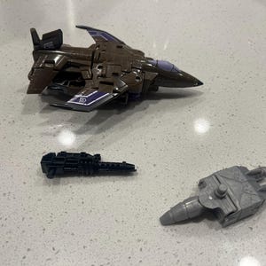 May include: A brown and purple toy jet with a black gun and a gray missile. The jet has purple accents and a detailed design. The gun is black and the missile is gray.