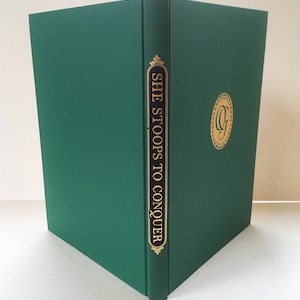 May include: A green hardcover book with gold lettering on the spine that reads "SHE STOOPS TO CONQUER". There is a gold circular design on the front cover with a letter "G" inside.