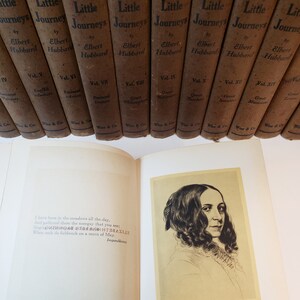 May include: An open book with a quote from the poem "Imparablene" by Emily Dickinson. The book is titled "Little Journeys" by Elbert Hubbard. The book is part of a set of books with the same title.