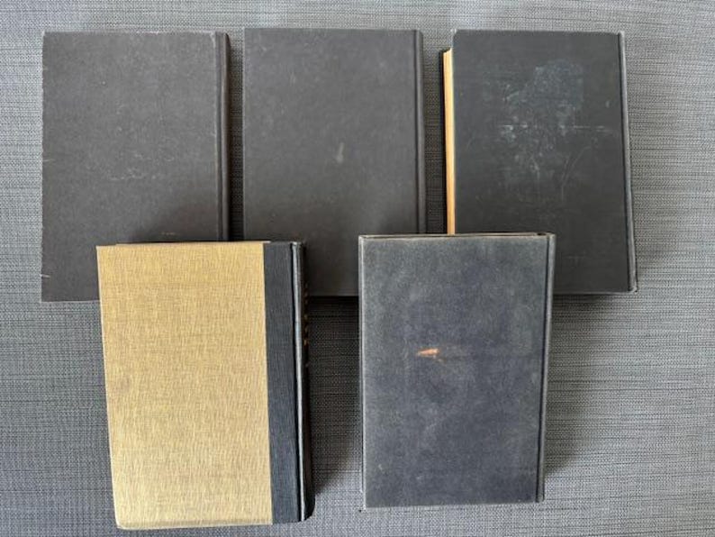 May include: A collection of five hardcover books. Four books have dark gray covers, while the fifth book has a gold cover with a black spine. The books are arranged on a gray surface.