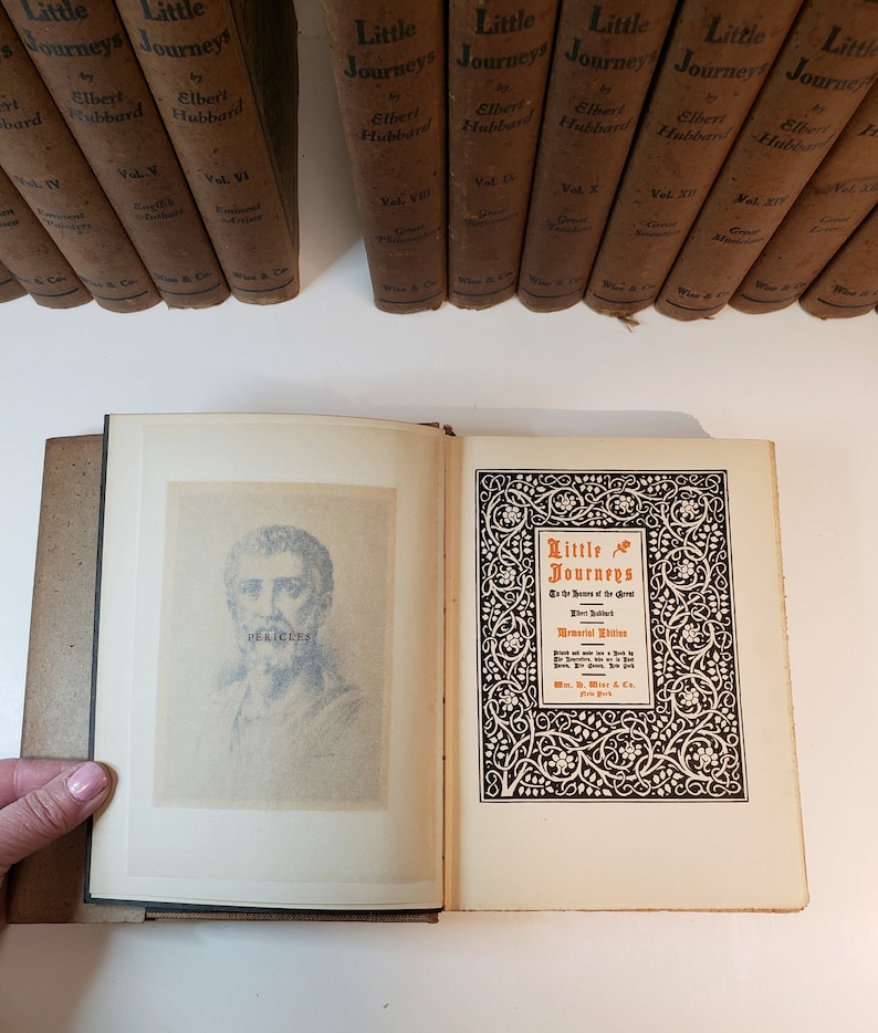 May include: An open book with a black and white illustration of Pericles on the left page and a title page for "Little Journeys to the Homes of Great Men" by Elbert Hubbard on the right page. The title page has a decorative border with a floral design.