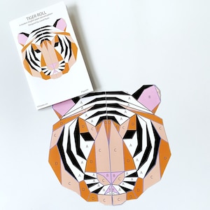 Pode incluir: Um padrão de cabeça de tigre em papel recortado com listras pretas e brancas, detalhes laranja e rosa e etiquetas de letras para cada peça. O padrão é intitulado "Tiger Roll" e foi desenhado por Laurel Ryan.