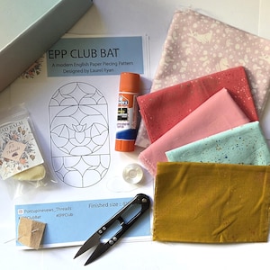 May include: An English Paper Piecing kit featuring a pattern sheet titled EPP CLUB BAT, a glue stick, a small clear bobbin, black thread snips, a needle in a felt square, a packet of thread balm, and five fabric swatches in pink, coral, and mustard.
