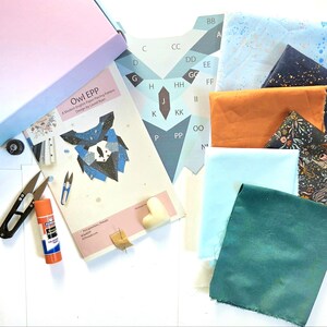 Könnte beinhalten: Eine Auswahl an Nähzubehör und Stoffstücken. Enthält ein "Owl EPP"-Muster, eine Schere, einen Klebestift und verschiedene Stoffmuster in Blau-, Orange- und Blumenmustern. Eine rosa Box ist in der oberen linken Ecke sichtbar.