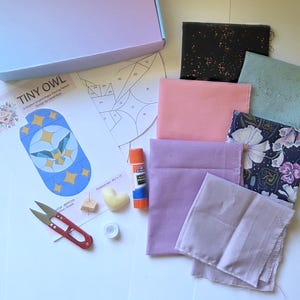 Könnte beinhalten: Ein Bastelset mit Stoffquadraten in verschiedenen Farben, darunter Rosa, Lavendel und Blumenmuster. Das Set enthält eine Papiervorlage, eine Schere, einen Klebestift und eine kleine herzförmige Kerze. Das Set ist für ein "Tiny Owl"-Projekt.