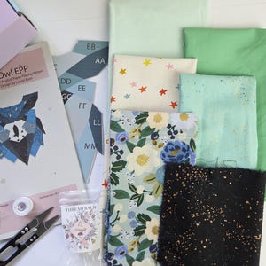 May include: A quilting kit featuring an Owl EPP English Paper Piecing pattern booklet, blue paper templates labeled AA through MM, a small spool of white thread, metal snips, a packet of Thread Balm, and assorted fabric swatches in green, floral, and black.