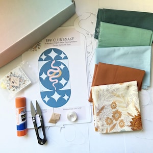 May include: An English paper piecing kit including a pattern sheet titled EPP CLUB SNAKE, a small container of thread balm, a glue stick, metal snips, a bobbin, and five fabric swatches in shades of dark green, light green, blue, and rust.