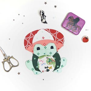 Può includere: Una collezione stravagante di materiali per l'artigianato e oggetti decorativi. Un'applicazione in tessuto a forma di rana con un cappello a fungo, una scatola di latta viola con un polpo, un bottone a forma di pomodoro, una statuina di puzzola e forbici dorate.