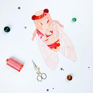 May include: A fabric cicada applique featuring a patchwork design with shades of pink, red, and gold, accented by a crescent moon pattern. The insect shape is constructed from multiple fabric segments with visible stitching, accompanied by a spool of pink thread and metal scissors.