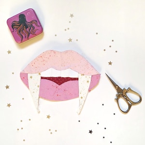 Puede incluir: Una boca de vampiro de tela rosa y blanca con detalles de estrellas doradas y largos colmillos. También hay una pequeña lata morada con un diseño de pulpo y unas tijeras doradas. Confeti de estrellas doradas esparcido.