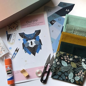 May include: An Owl EPP English Paper Piecing pattern by Laurel Ryan, including a stack of folded fabric in green, mustard, and floral prints, a glue stick, thread balm, metal snips, a bobbin, and a paper template sheet labeled with letters.