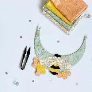 May include: A bee-shaped fabric applique with a crescent moon background. The bee is made of yellow, black, and white fabric, and the moon is made of light blue fabric with gold dots. The applique is surrounded by small gold stars.