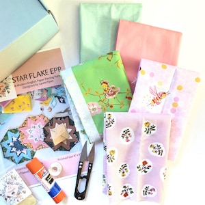 May include: A quilting kit featuring a Star Flake EPP pattern booklet, a light blue box, a glue stick, metal snips, and several folded fabric pieces in shades of green, pink, and lavender with bee, honeycomb, and floral prints.