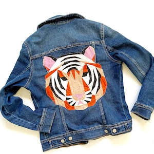 May include: A blue denim jacket featuring a large, geometric tiger head appliqué on the back. The tiger design is constructed from multi-colored fabric pieces in shades of orange, white, black, and pink, showcasing a stylized, angular face.