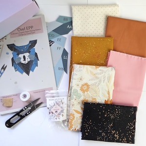 Könnte beinhalten: Ein Owl EPP-Anleitungsheft für englisches Papierschablonen-Patchwork, blaue Papierschablonen mit der Beschriftung AA bis MM sowie sechs Stoffmuster in Creme, Rost, Senfgelb, Rosa, Blumenmuster und Schwarz mit Goldsprenkeln, plus Fadenschere.