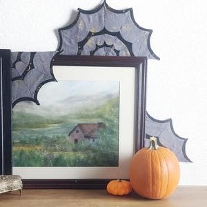 May include: A framed landscape painting of a house near a lake, decorated with grey and black fabric spider web corner accents featuring gold celestial patterns. Two orange pumpkins sit in front of the dark brown wooden picture frame.