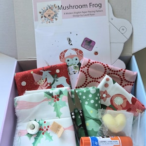 Könnte beinhalten: Ein Bastelset in einer Schachtel mit einer Mushroom Frog Patchwork-Anleitung, verschiedenen gemusterten Stoffstücken in Rot, Grün und Weiß, einer Metallschere, einer weißen Spule, einem kleinen Filzstück, einem herzförmigen Teil und einem Klebestift.