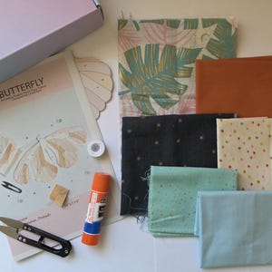 Könnte beinhalten: Ein Bastelset mit verschiedenen Stoffen, einem Schmetterlingsmuster, einer Schere, einem Klebestift und Faden. Die Stoffe umfassen Drucke mit Blättern, Sternen und unifarbenen Farben. Das Set ist für Nähprojekte konzipiert.