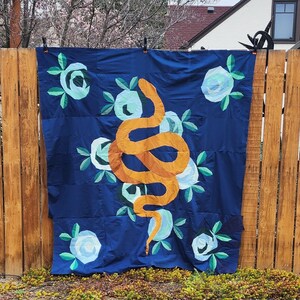 Op de afbeelding: Een donkerblauwe quilt met een oranje slangenontwerp en witte en lichtblauwe bloemaccenten. De quilt hangt aan een houten hek. De slang is de centrale focus van de quilt.