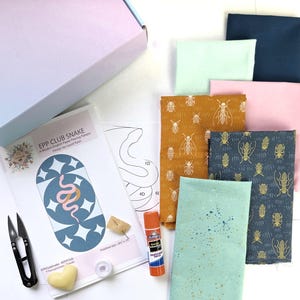 Könnte beinhalten: Ein Bastelset mit Stoffquadraten in verschiedenen Farben und Insektenmustern, einer Papiervorlage, einer Schere, Klebstoff und anderem Zubehör. Das Muster trägt den Titel "EPP CLUB SNAKE". Fertige Größe: 21,6 x 38,1 cm.