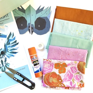 Könnte beinhalten: Ein Quilt-Set mit einer Papiervorlage für eine Eule, einem Stapel aus fünf Stoffstücken in Orange, Hellblau, Grün, Rosa und Blumenmustern, einem Klebestift, einer kleinen Spule mit weißem Garn und einer schwarzen Metall-Fadenschere.